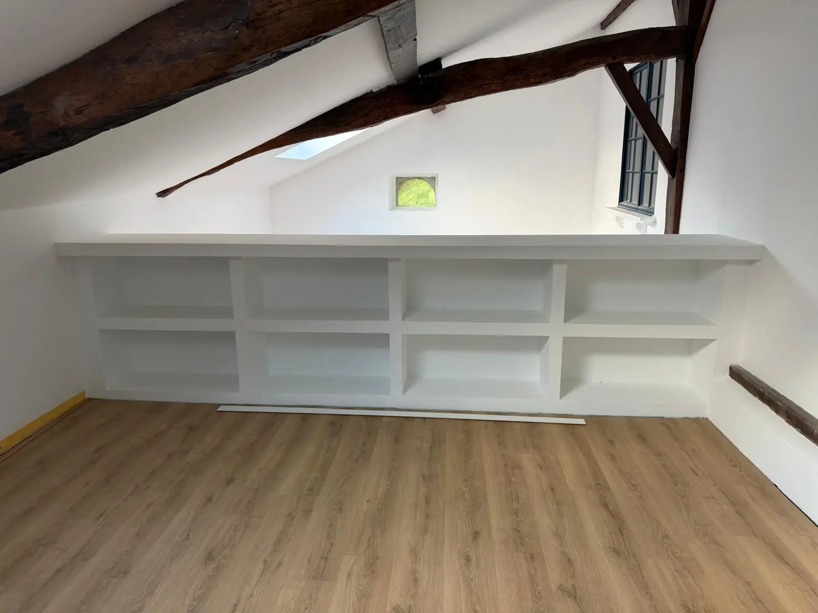 Bibliothèque basse en plaques de plâtre peinte en blanc, sous un rampant aux poutres anciennes, parquet
